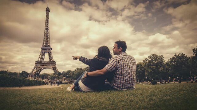 Romantic couple sitting together on grass looking at the Eiffel Tower in Paris under a cloudy sepia-toned sky.
