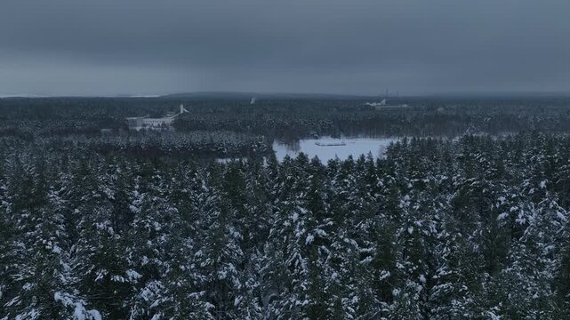 Drone descends and pans over snow covered pines in Belarus, revealing a frozen reservoir, a small dam, a pale building with tower, and distant steaming chimneys.