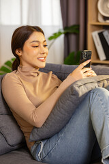 Obraz premium Happy asian woman relaxing on comfortable sofa holding smartphone enjoy watching funny video content online social media feeling cheerful smile at home living room apartment