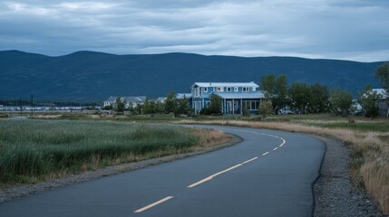 Fototapeta premium Winding asphalt pathway leads toward clustered residential structures backed by dark mountain range under overcast sky