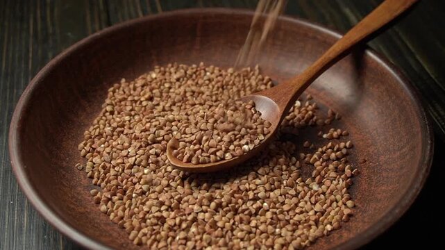 Brown seeds buckwheat being poured from a wooden spoon into a bowl filled with more seeds, showcasing the continuous flow of the action in a rustic kitchen setting