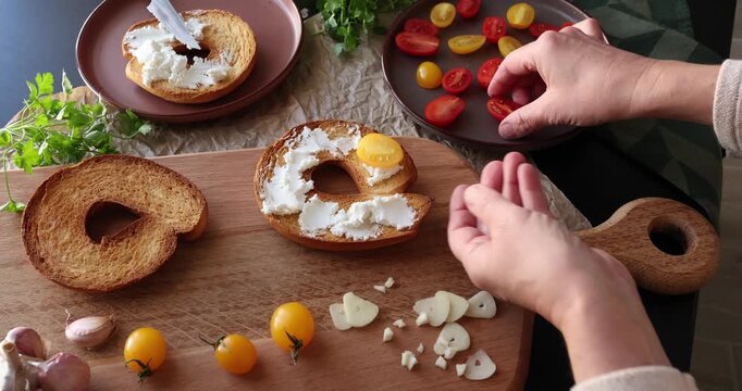 Anonymous person prepares round bruschetta with creamy goat cheese and cherry tomatoes in kitchen. Italian cuisine, high-quality farm-grown ingredients.