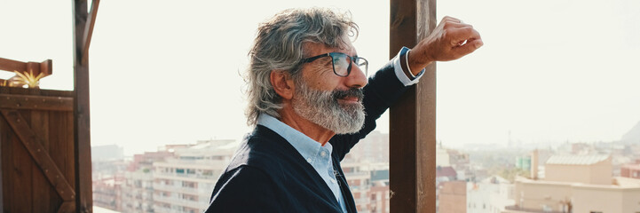 Fototapeta premium Senior enjoys the view standing on the balcony with mobile phone in hand. Panoramic