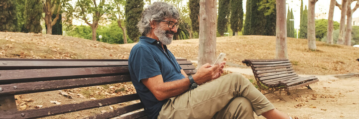 Smiling elderly man in glasses using phone while sitting on park bench. Panoramic. Panoramic © Andrii Nekrasov