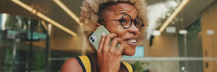 Close up, happy laughing young woman in glasses, wearing brown top, with backpack on her shoulders, stands on the street and emotionally talks on mobile phone. Panoramic. Panoramic © Andrii Nekrasov