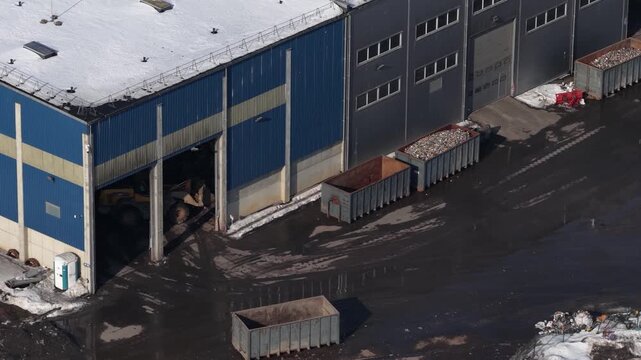 Drone view of recycling facility yard with front-end loader, large metal waste containers and scattered debris on wet asphalt, surrounded by snow in winter.