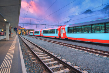 Obraz premium Red and blue modern passenger train standing at railway station in Swiss Alps at dusk. Mountains in fog, tracks and railway platform under purple cloudy sky. St. Moritz, Switzerland. High-speed train