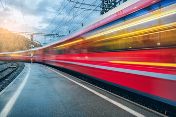 Fototapeta premium Blurred red passenger train passing mountain railway station during rain in Swiss Alps at sunset. Moving speed train at dusk. St. Moritz, Switzerland. Bernina Express. Railway platform and lights.