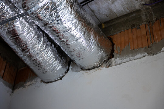 Insulated ventilation ducts passing through wall opening during house construction. HVAC installation detail with foil air pipes in unfinished interior before ceiling completion.