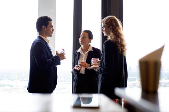 Business professionals networking by a large window at a corporate event, discussing strategy and partnership while holding drinks in a modern office setting with city view background