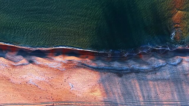 Aerial perspective of ocean waves hitting sandy beach at sunset. Horizontal drone shot showing sea surf meeting a sandy shore with long shadows and footprints.