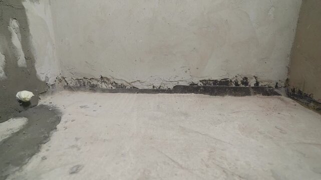 Close-up of an unfinished room corner during construction. Raw concrete floor plastered wall meet with a black waterproofing membrane. Industrial texture neutral gray colors. Work in progress.