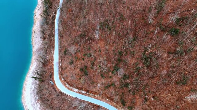 Top down aerial view of asphalt road by blue lake. Curving road separates turquoise water from a forest of bare trees with orange and brown leaves.