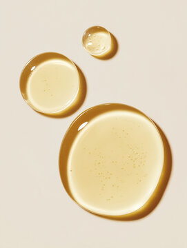 Top view macro image of three glossy cosmetic oil drops on a clean light beige background. The viscous transparent serum has a natural golden tone and smooth texture, suitable for skincare, beauty, sp