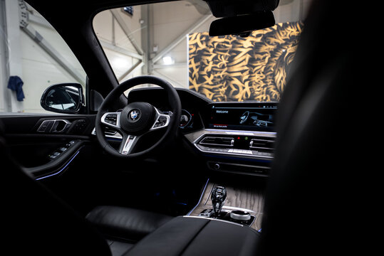 Driver's seat view of a white BMW X7 cabin shows welcome screen, leather wheel with BMW roundel, crystal gear selector, iDrive controller, ambient accents, and mural.