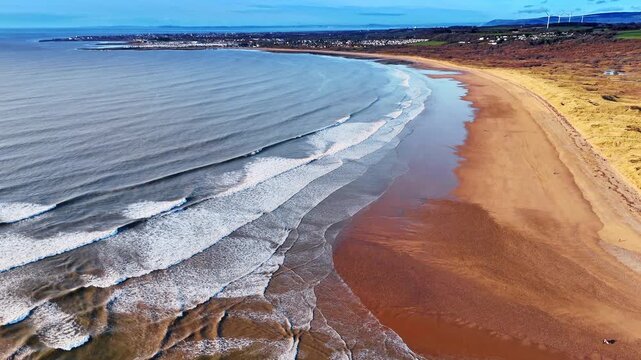 Long coastline with rolling waves and wide sandy shore. High altitude view of parallel wave lines approaching a vast sandy beach.