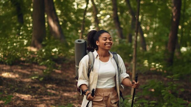 A young African American woman enjoys her time hiking in a forest. She carries a backpack and hiking poles while smiling. The scene is filled with green trees and nature around her.