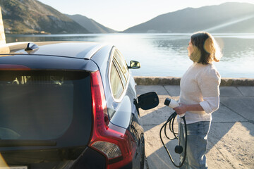 Woman traveler with charging plug next to her electric car on the sea coast at sunset