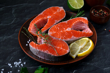 Raw trout steaks, lemon wedges, rosemary, and thyme on a brown plate. Peppercorns, coarse salt, lime, and parsley surround them against a dark background. Fresh seafood