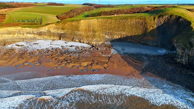 Aerial view of stratified limestone cliffs and rocky beach. Direct drone shot showing geological layers of cliffs and a pebble beach with scattered rocks at low tide.