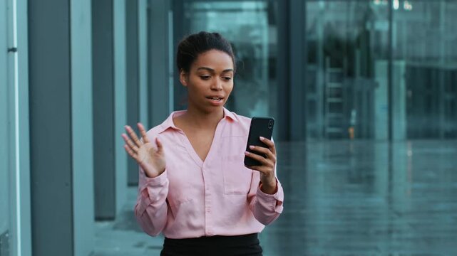 A woman with dark skin stands indoors in a modern building, looking at her smartphone with a joyful expression. She is dressed in a light pink blouse and black skirt.
