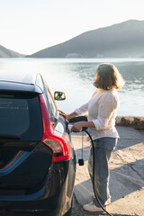 Fototapeta premium Woman traveler with charging plug next to her electric car on the sea coast at sunset