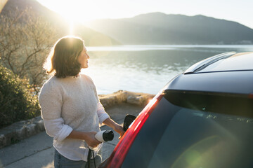Fototapeta premium Woman traveler with charging plug next to her electric car on the sea coast at sunset