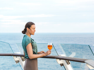 Stylish woman with a glass of drink standing on the empty deck of a cruise ship against the backdrop of sea waves. Sunny day. Chill lifestyle, luxury travel. Perfect for holiday and travel themes © Svetlana