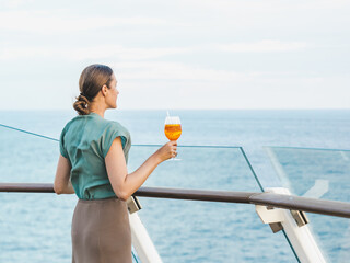 Stylish woman with a glass of drink standing on the empty deck of a cruise ship against the backdrop of sea waves. Sunny day. Chill lifestyle, luxury travel. Perfect for holiday and travel themes © Svetlana