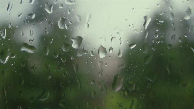 Raindrops hit and slide down a windowpane. The background is blurred due to shallow depth of field. The focus is on the clear water droplets forming patterns on the glass.