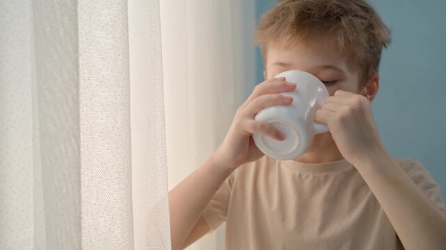 Boy at the window drinking milk, tea or cocoa from a white ceramic mug. Focus on child nutrition, dairy products, breakfast routine, and healthy beverage intake at home for kids.