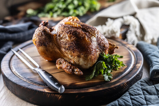 A whole chicken, baked until golden brown, is served on a wooden cutting board, garnished with fresh parsley and accompanied by a meat fork in a rustic setting