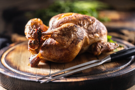 A whole chicken, baked until golden brown, is served on a wooden cutting board, garnished with fresh parsley and accompanied by a meat fork in a rustic setting