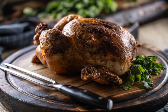 A whole chicken, baked until golden brown, is served on a wooden cutting board, garnished with fresh parsley and accompanied by a meat fork in a rustic setting