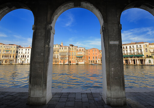 Scenic view of the Grand Canal in Venice framed by historic stone arches. Colorful Venetian palaces reflected in the water under blue sky. Iconic Italian architecture, travel destination and romantic 