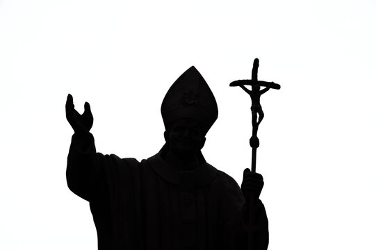 A Catholic pastor in a cassock and cap with a cross in his hand, silhouetted against a light background.