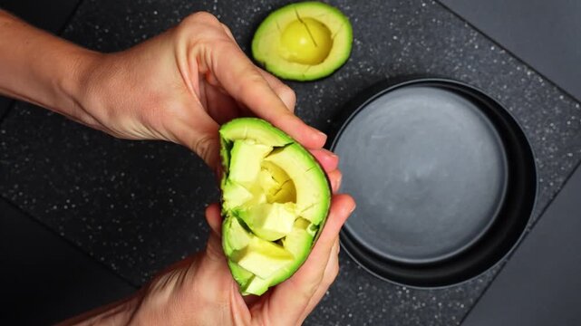 Hands holding halved avocado with cut flesh over bowl. Close-up of person squeezing an avocado half to release diced fruit pieces into a black container.