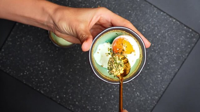 Hand spooning furikake seasoning over soft boiled egg bowl. Close-up of person adding spices to a cooked egg in a small green ceramic bowl using a gold spoon.