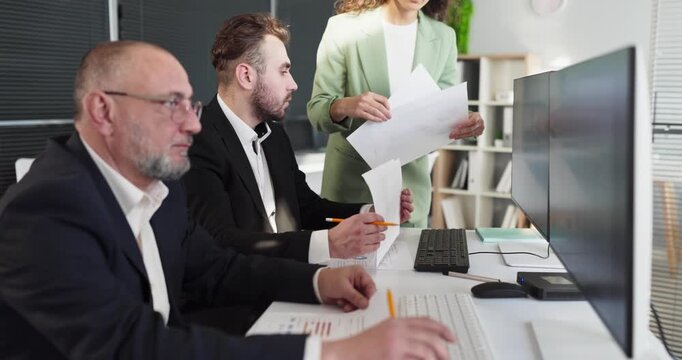 Analysts team in office reviewing finance data. Colleagues at computers compare report data, track statistics, check paperwork, align budgets in a meeting. Strong teamwork and analytical mindset.