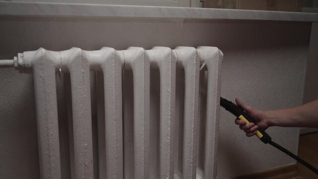 Person hand holding steam cleaner nozzle cleaning white cast iron heating radiator, ensuring hygiene and sanitation in home environment. Concept of maintenance, hygiene, routine indoor cleaning.