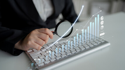 A business professional inspects financial data through a magnifying glass on a keyboard,...
