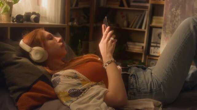 Side view shot of red haired young woman listening to calming music and wearing big headphones while napping on couch at home playing playlist in mobile app