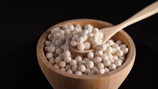 White tapioca pearls in wooden bowl spinning over black background. Raw cassava starch balls used for cooking and bubble tea. White dry tapioca pearls in wooden bowl with spoon.