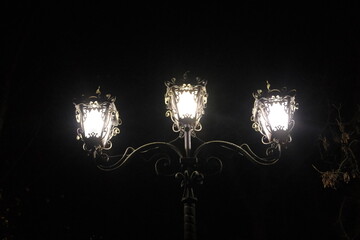 Detailed lamppost with three illuminated lanterns stands tall, casting a bright light into the dark night. © vannet