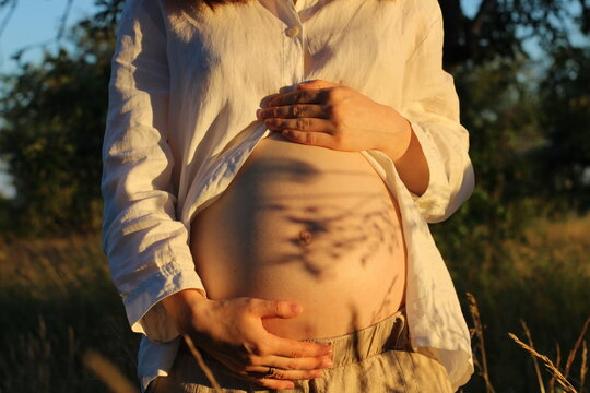 Exspecting mother gently holding baby bumb in soft sunlight with plant shadows