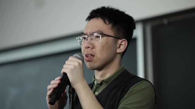 A young Asian male wearing glasses shows a nervous, anxious expression while speaking into a handheld microphone, ideal for fear of public speaking, stage fright, or student anxiety concepts.