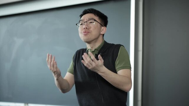 A young Asian male wearing glasses gestures expressively while reciting a monologue in a classroom setting, ideal for drama class, acting performance, or performing arts education concepts.