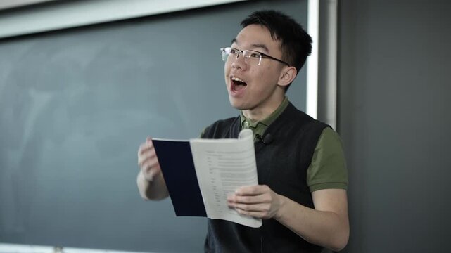A young Asian male wearing glasses reads from a script book while performing in a classroom setting, ideal for drama class, script reading, or performing arts education concepts.