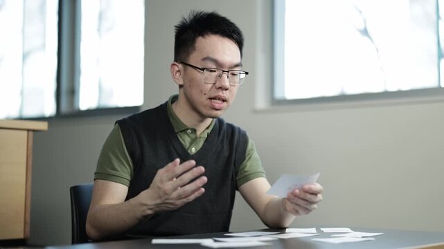 A young Asian male student wearing glasses reads and pronounces words from flashcards at a desk, ideal for language learning, vocabulary study, or foreign language education concepts.