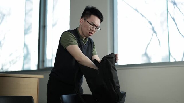A young Asian male student wearing glasses rummages through his backpack and scratches his head unable to find something, ideal for forgetfulness, student life, or everyday frustration concepts.